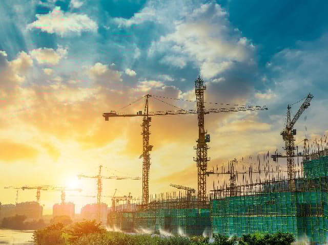 Construction site worker with hard hat and surveying equipment looking up at crane and preparing for building materials inspection