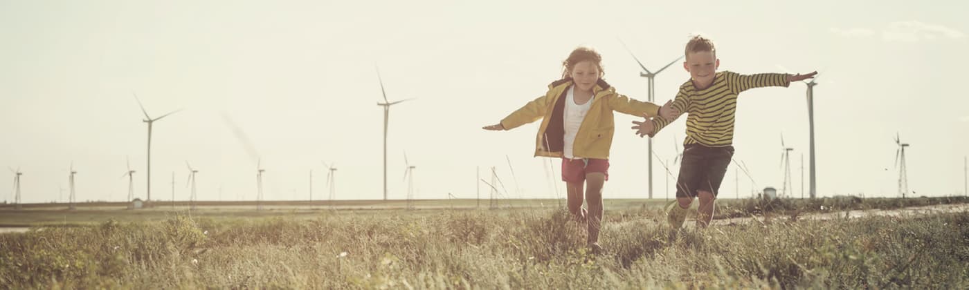 Two children running in an open field of long grass with windmills in the sky behind them