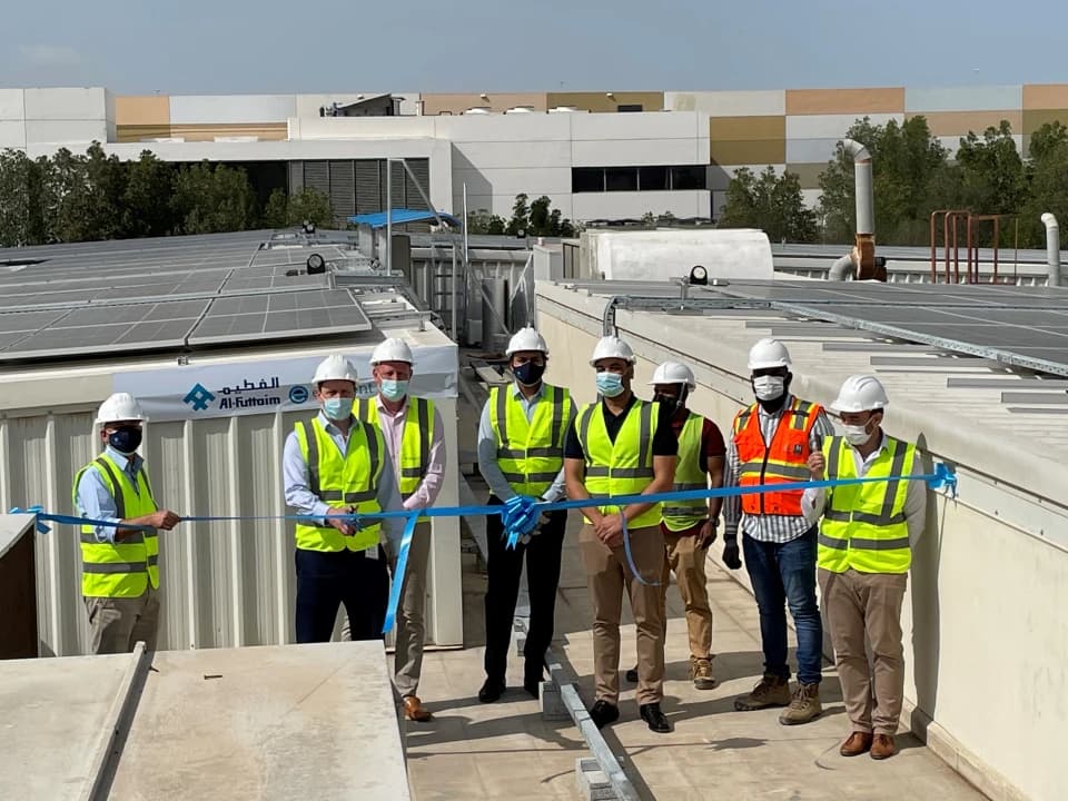 Element colleagues standing on the roof of one of their UAE labs, wearing green work vests and helmets, cutting a ribbon with their newly installed solar panels in the background
