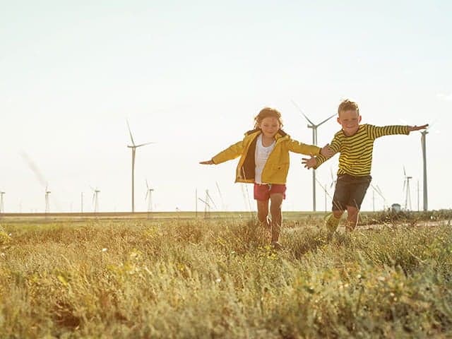 Two children running in an open field of long grass with windmills in the sky behind them