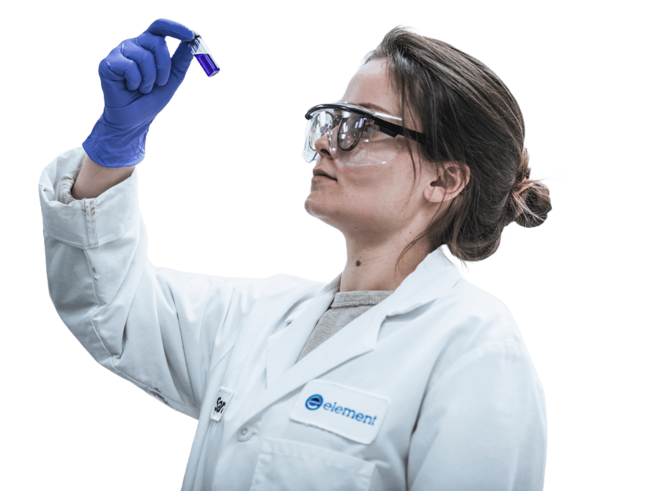 Element laboratory scientist wearing safety goggles and a white lab coat with the Element logo, inspecting a test tube containing a purple sample during materials testing.