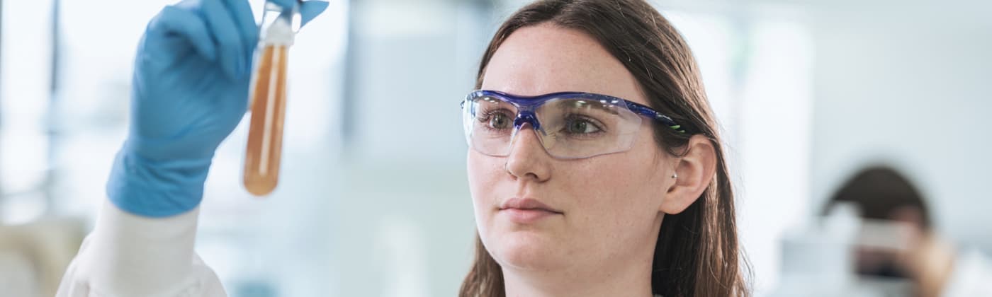 Female scientist in lab coat and safety glasses examining test tube containing amber-colored biological sample