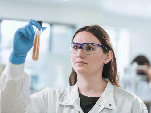 Female scientist in lab coat and safety glasses examining test tube containing amber-colored biological sample