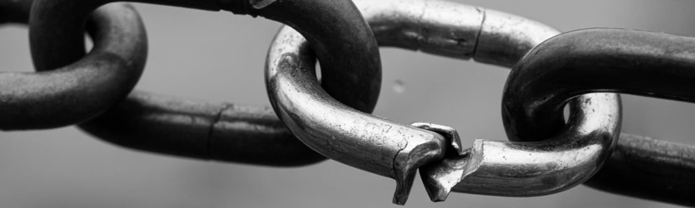 Close-up black-and-white image of a metal chain with one link snapped apart, showing rough, jagged edges where the break has occurred against a blurred background.