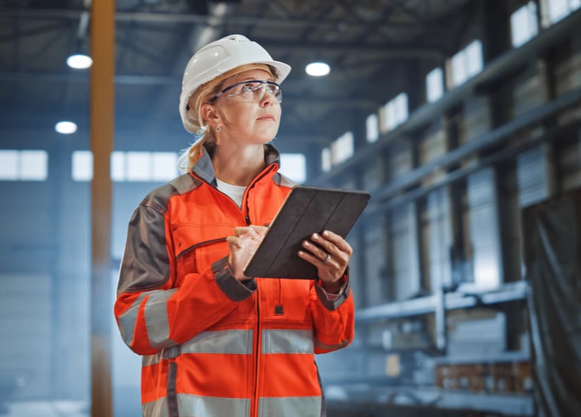 Woman in hard hat and orange work jacket entering information on tablet at a construction site
