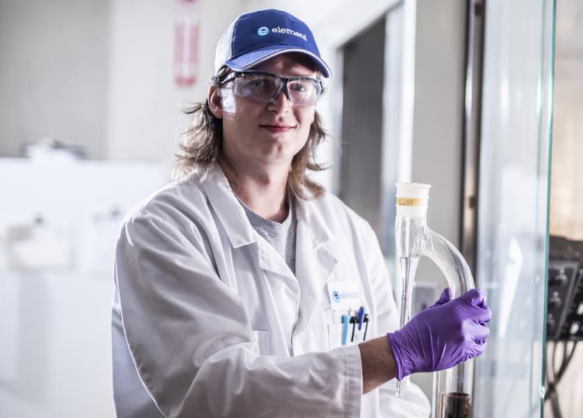 Element scientist with white coat and baseball hat standing in lab with large chemical analysis container