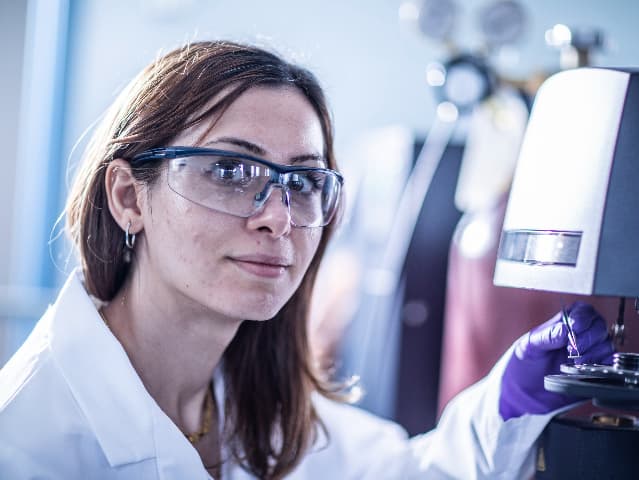 Scientist wearing safety glasses and gloves looking at camera and adjusting a testing machine next to her